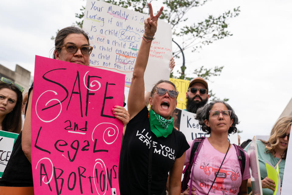 A protester yells and points two fingers in the air, with a woman at her side carrying a poster that says: Safe and Legal Abortions, and one behind her saying: Your Morality is ... lacking if all you want is a child Born. But not a child Fed. Not a child educated. Not a child Housed. That's not Pro-Life. 