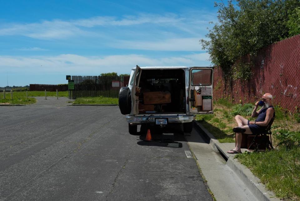 A man sits outside a panel van with its rear doors open, with a landfill just beyond where he parked