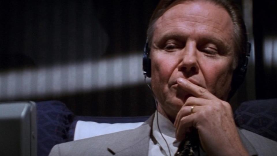 Jon Voight listens to headphones while wearing a suit on a plane in Mission: Impossible