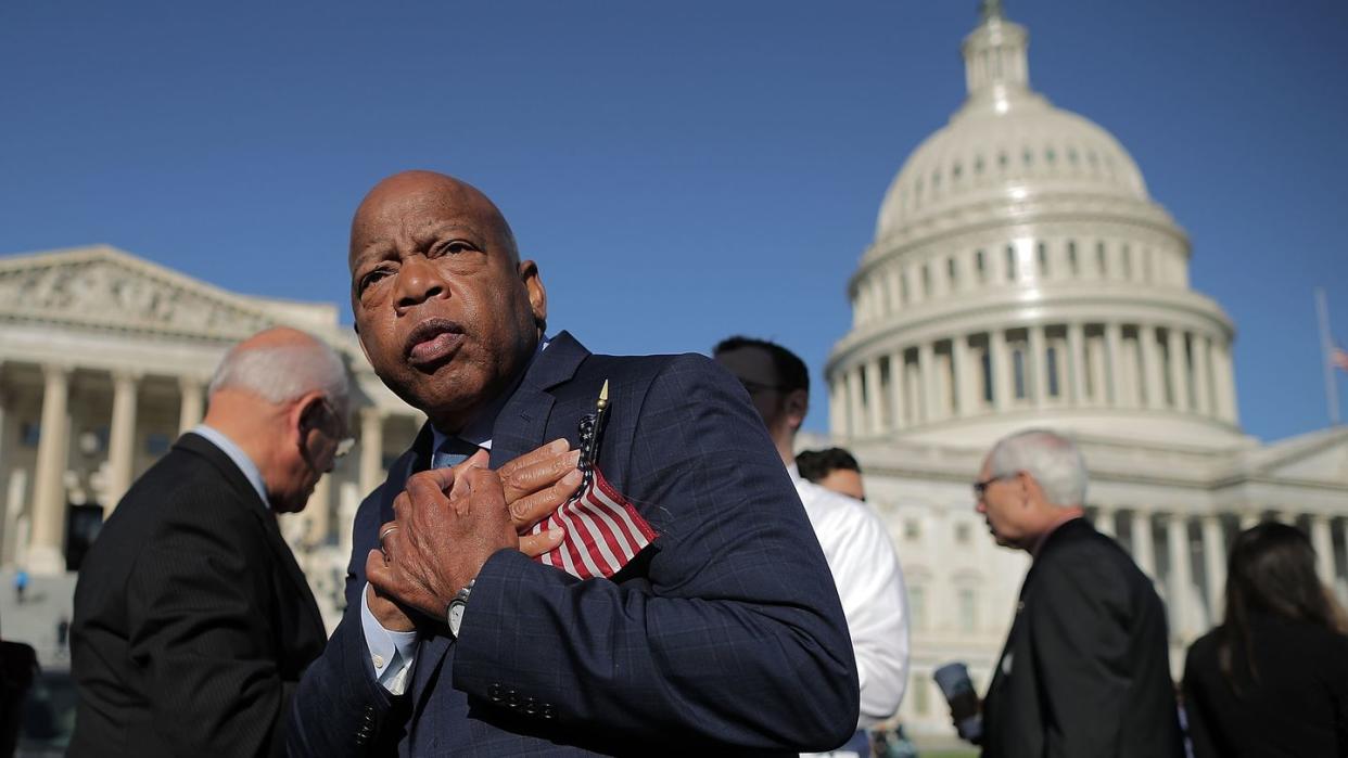 congressman john lewis holds a small american flag to his chest, he stands outside the us capitol building in a suit jacket, other people stand in the background