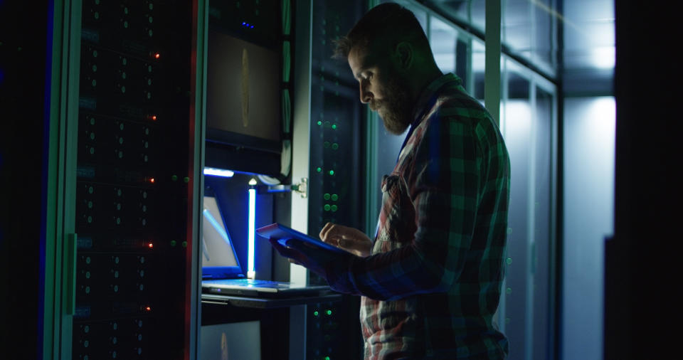 Side view of modern IT engineer using tablet and laptop setting servers in data center while working on mining farm