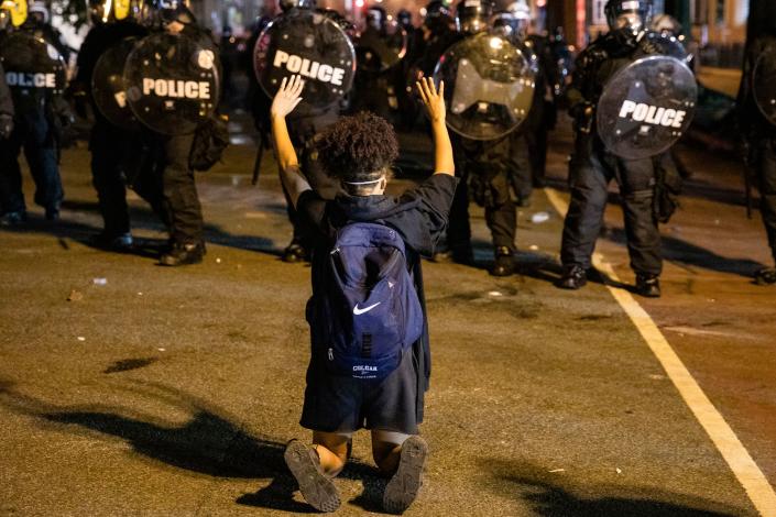 A demonstrator kneels and raises her hands facing a  police line