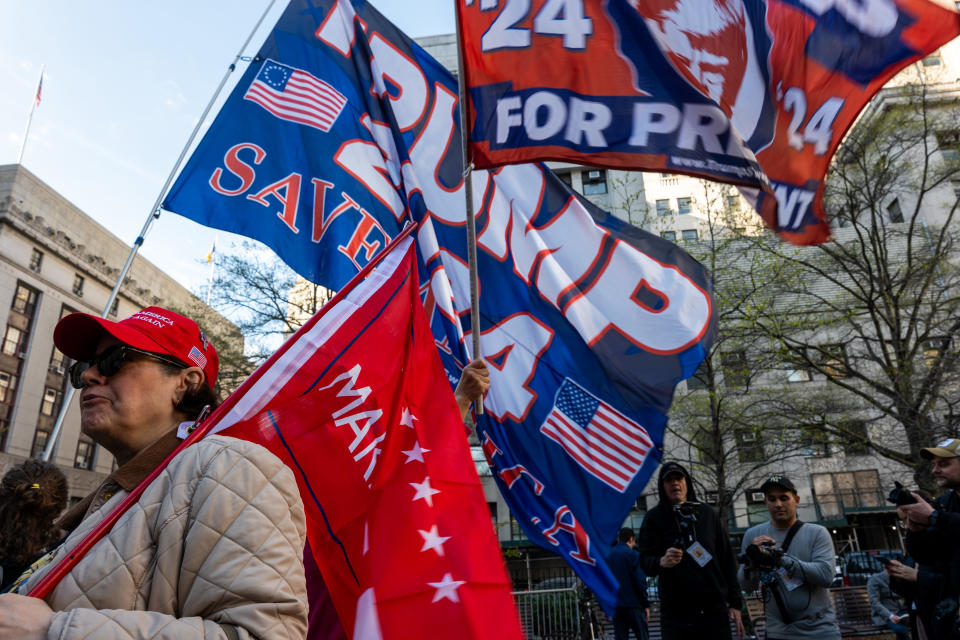 Supporters with "Trump 2024" flags at a rally, one wearing a red hat