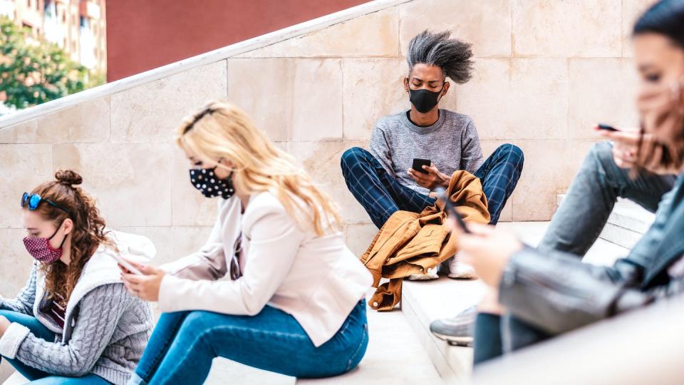 Students sit on university steps looking at smartphones and wearing masks.