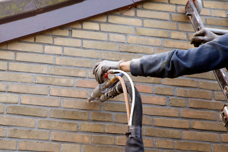 A worker up a ladder injects insulating foam into a brick wall.