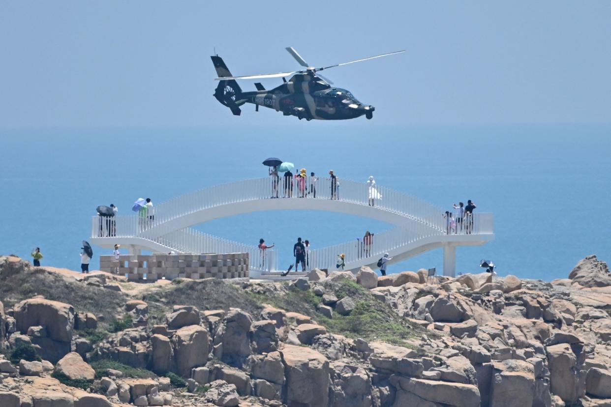 People with sun umbrellas stand on an arched, elevated walkway along a rocky coast facing a flying helicopter. 