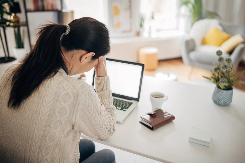 Woman seated at a desk with a laptop, appearing stressed or thoughtful, with a coffee mug beside her