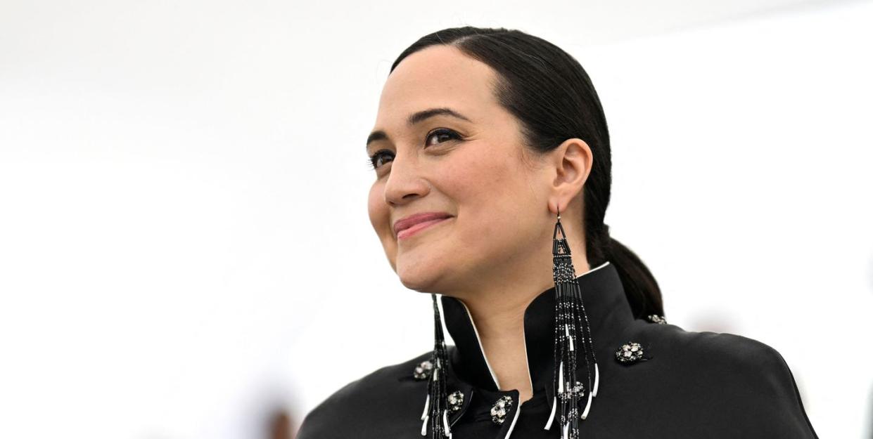 a closeup of lily gladstone posing for photographers at cannes film festival, she's wearing her hair in a ponytail and is rocking a black cape dress adorned with rhinestone buttons and accessorised with black chandelier earrings