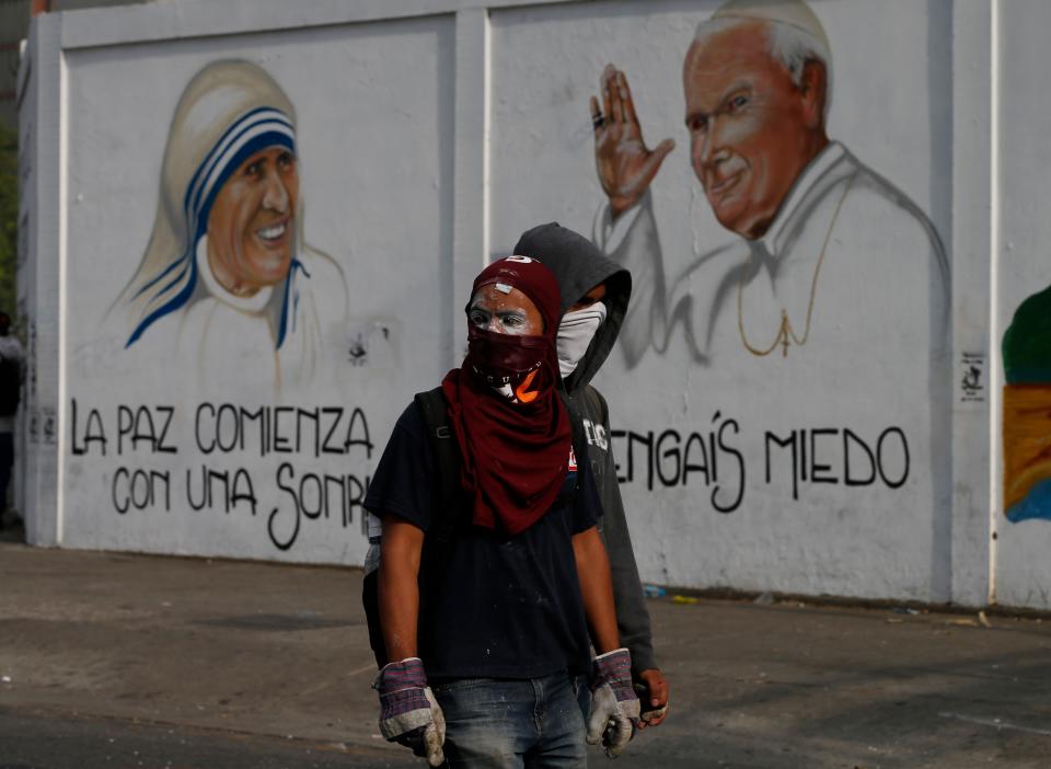 Two masked demonstrators walk in front of a mural with painted images of Mother Teresa and Pope John Paul II during clashes with Bolivarian National Police in Caracas, Venezuela, Tuesday, April 1, 2014. Protesters had gathered to march to the National Assembly with opposition lawmaker Maria Corina Machado, who was stripped of her parliamentary seat last week, after addressing the Organization of American States about the conditions of Venezuela. Riot police prevented the protesters from reaching the parliament building. (AP Photo/Fernando Llano)