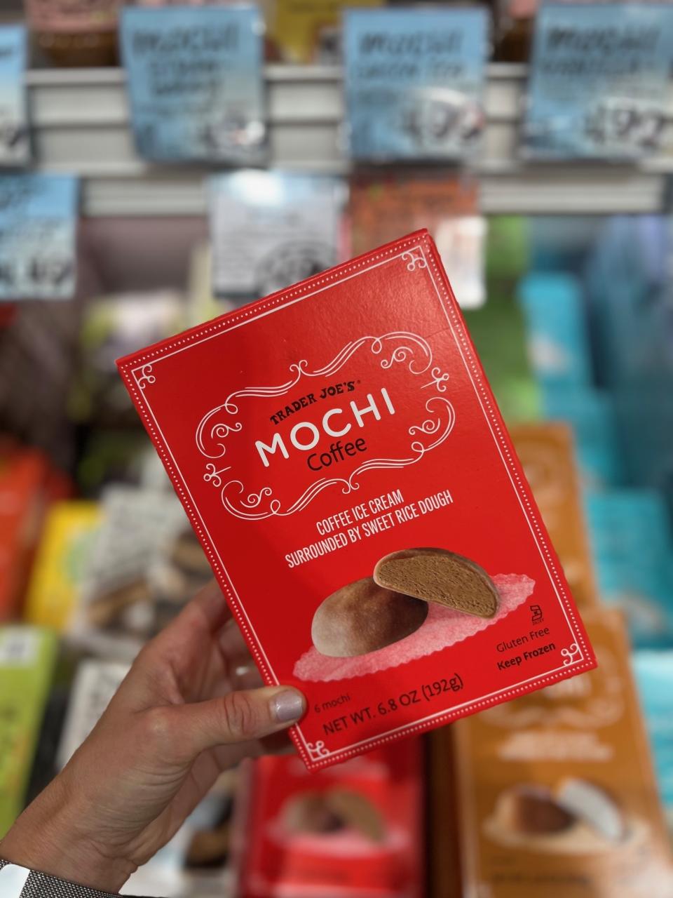 A hand holding a box of Trader Joe's Mochi Coffee ice cream. The box has text detailing the product as "coffee ice cream surrounded by sweet rice dough."