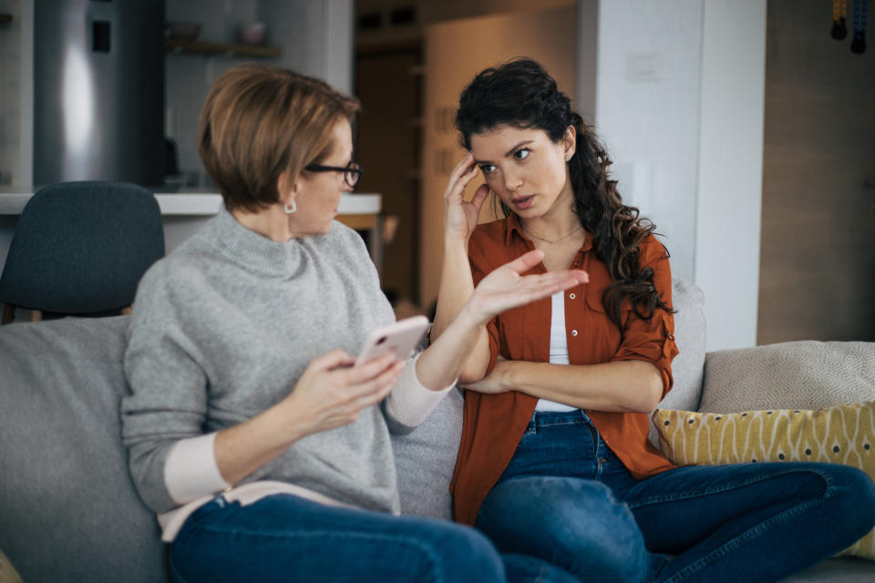 Two women, one older with glasses holding a phone, and one younger with curly hair, sitting on a couch and engaged in a serious conversation at home