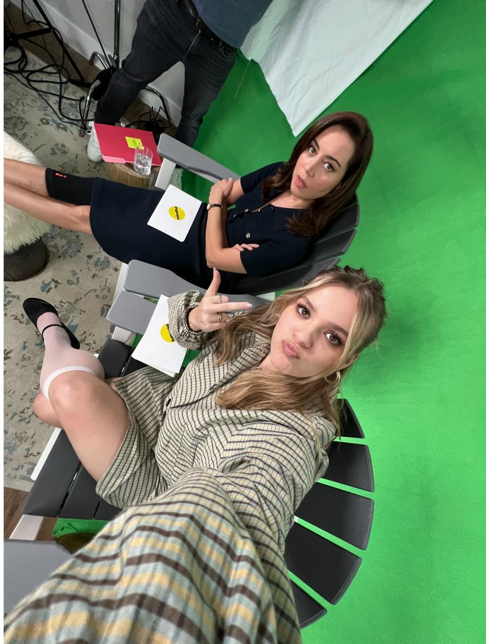 Aubrey Plaza and Maisy Stella sit together on a set with a green screen background, posing playfully for the camera. They appear casual and relaxed