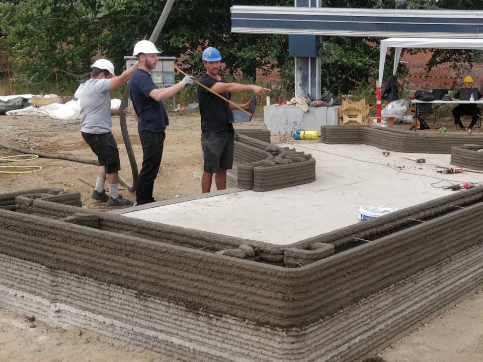 Workers in hard hats pointing to a short 3D printed wall.