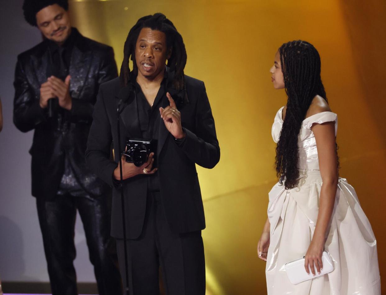 Jay-Z with dreads in a black suit standing on a stage next to Blue Ivy in a white dress.