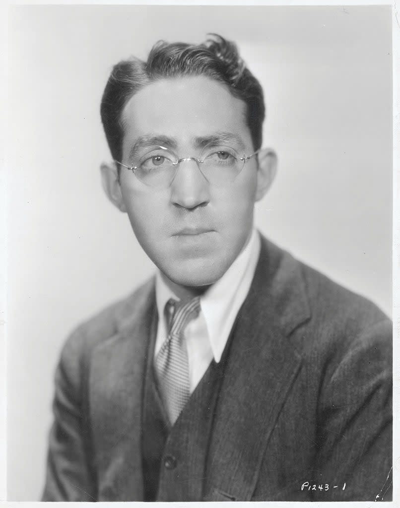 A man in a suit and tie with glasses, posing for a formal portrait. His hair is styled neatly, and he has a serious expression. Names are not provided