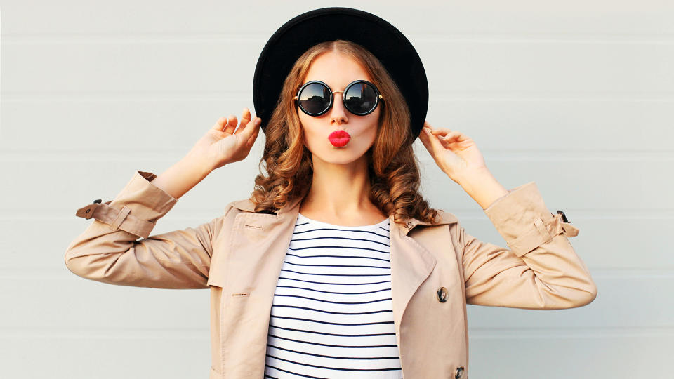 Fashion portrait pretty sweet young woman blowing red lips wearing a black hat sunglasses coat over grey background.