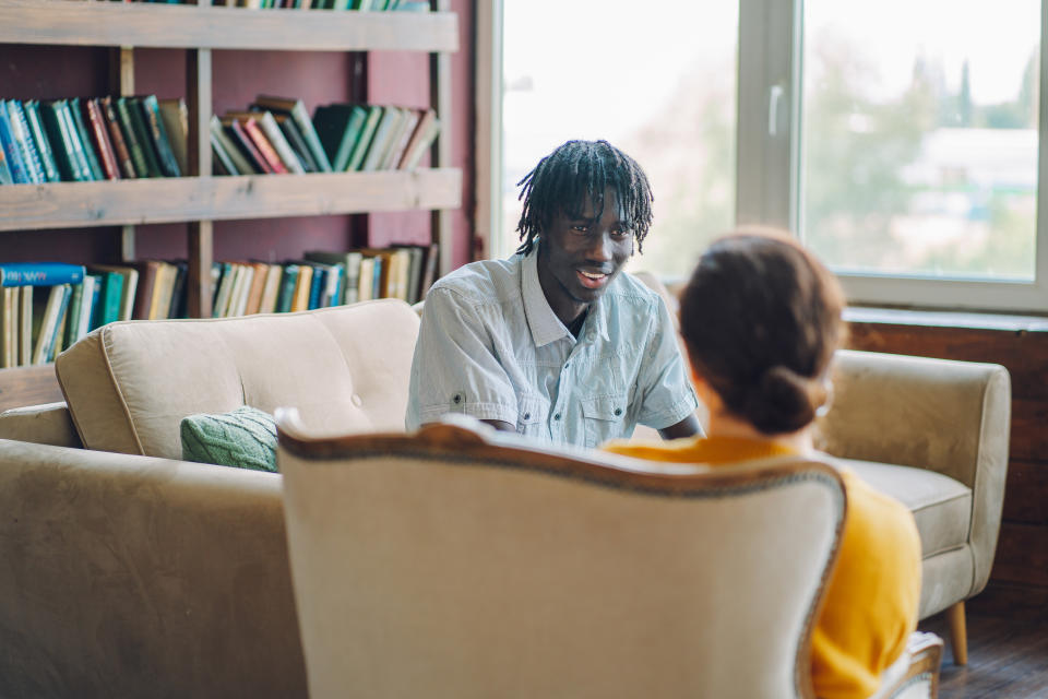 A man and woman are having a conversation on a couch in a cozy room filled with bookshelves