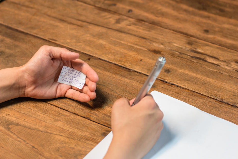 Hand holding a small paper with scribbled notes while writing on a blank sheet, suggesting use of a cheat sheet