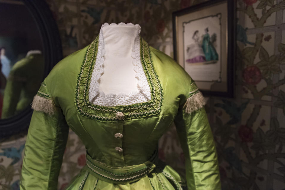 Antique green 19th-century dress exhibited in a museum, adorned with intricate lace details on the neckline and fringed trim on the shoulders