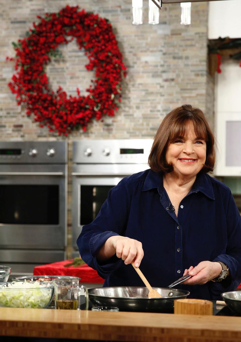 Ina Garten in a kitchen, smiling and stirring something in a pan with a wooden spoon.