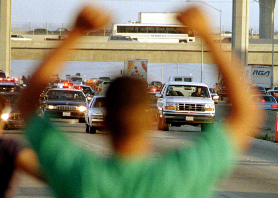 A man on the side of the road appears to cheer as a Ford Bronco drives along a highway followed by police cars.