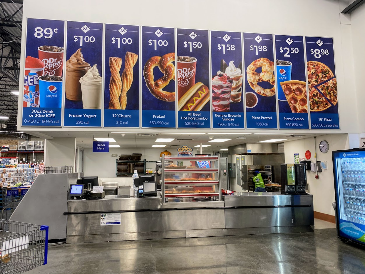 Cafe counter at a Sam's Club, large photos with pricing on the top, clean area and concrete floor, warehouse in background on the left