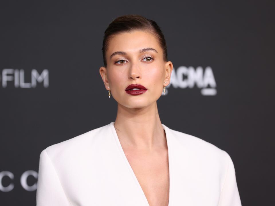 Hailey Bieber at an event in a white dress and dark red lipstick.