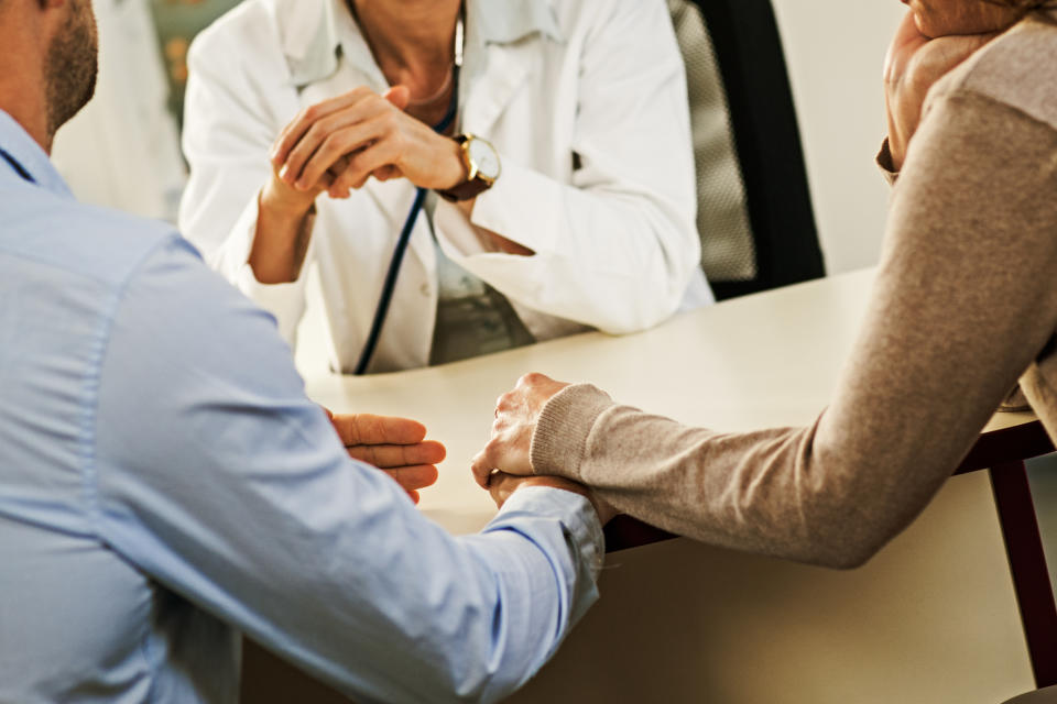 Two people holding hands across a table, with another person watching, in a supportive setting