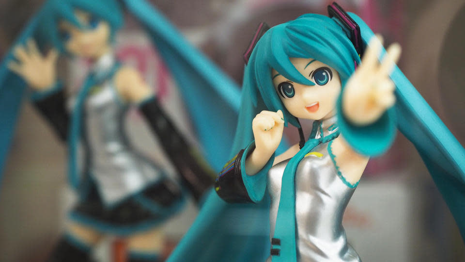 Primer plano de una figura de Hatsune Miku.  Ella agita un signo de la paz hacia la cámara mientras una segunda figura en una pose diferente se sienta (borrosa) detrás.