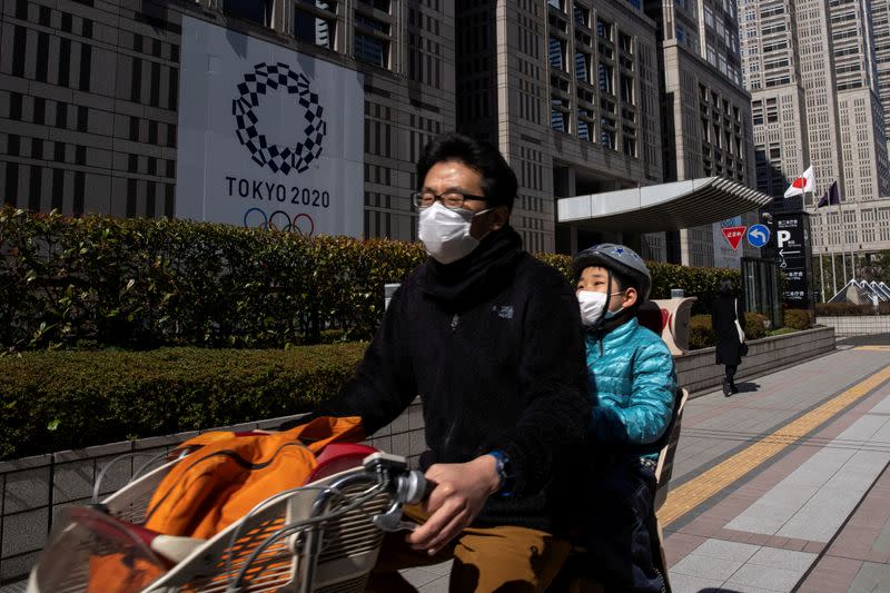 FILE PHOTO: A father and son wearing protective face masks ride a bicycle past a banner for the upcoming Tokyo 2020 Olympics
