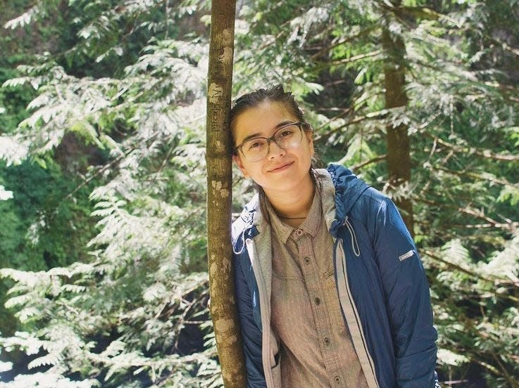 The author leans against a thin tree with more trees behind her