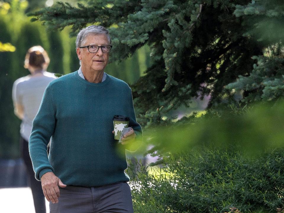 Bill Gates holding coffee cup walking among pine trees at Sun Valley conference