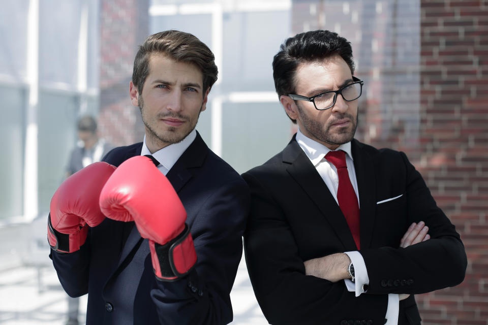 Two businessmen standing back to back in an alley. One is wearing red boxing gloves.