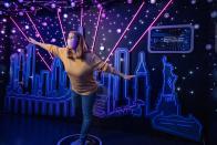 <p>Google Store Chelsea. A Night Sight-themed Sandbox with a person wearing a face mask posing in the middle. The background features a cut-out art rendering of the New York City skyline with rays of pink light emanating from it and the Statue of Liberty on the right. A display is on the wall above this. </p> 