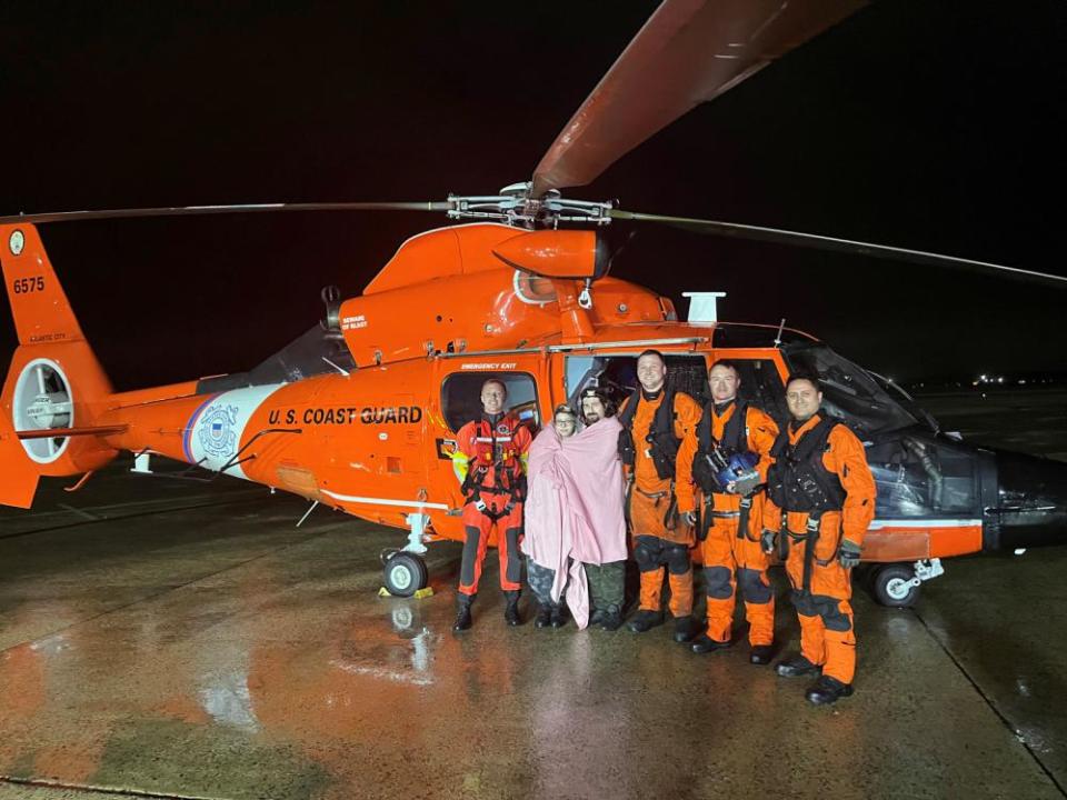 An MH-65 Dolphin helicopter crew from Coast Guard Air Station Atlantic City stands with two survivors May 6, 2022, after the aircrew rescued the couple. The two people were aboard a 33-foot sailing vessel when it grounded and overturned in heavy weather offshore from Little Egg Inlet,