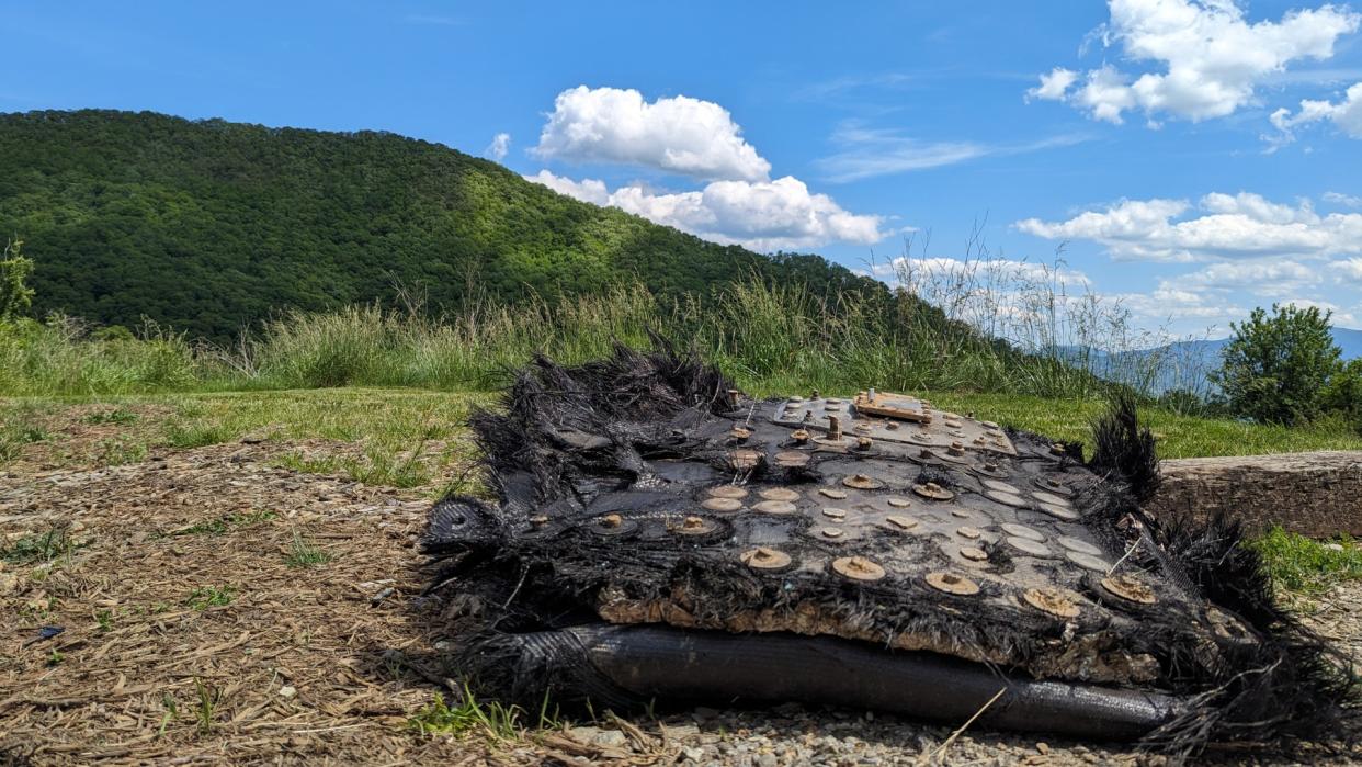  A large black piece of fiberglass covered in metal bolts and plates lies on the ground beside a trail leading into a forest. mountains can be seen rolling in the distance. 