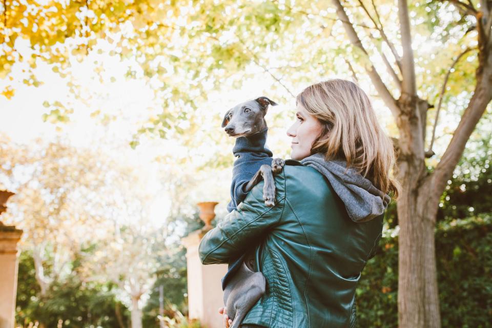 woman wearing a leather jacket holding an italian greyhound wearing a turtleneck sweater outdoors