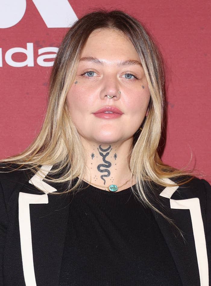 Elle King at a red carpet event, wearing a dark outfit with a white-trimmed blazer. She has multiple face and neck tattoos