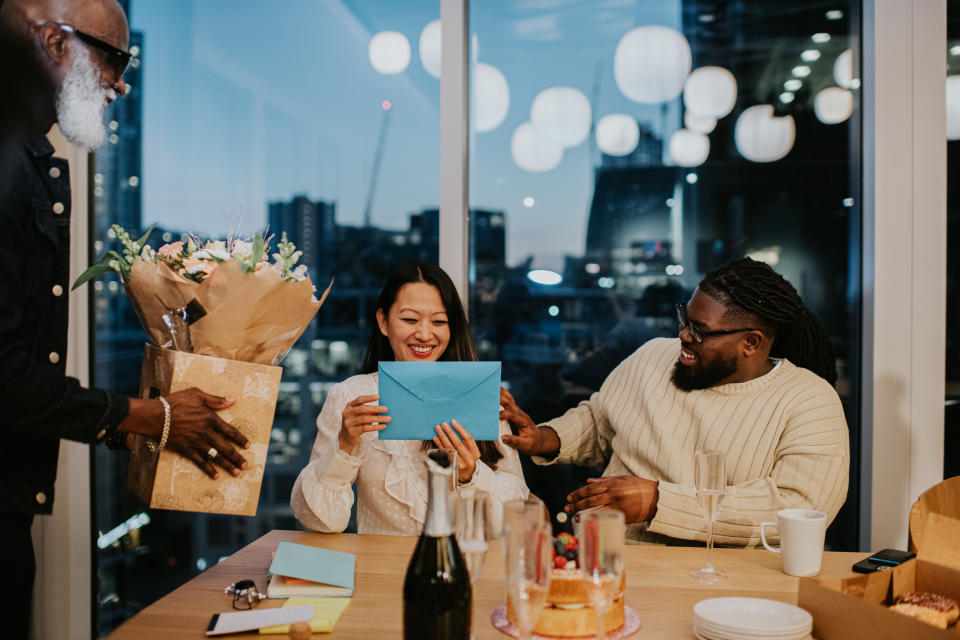 An after-work office party for a female employee. Colleagues lavish attention and praise on her, presenting her with a bouquet of flowers and a card. Champagne and a cake sits on the table.