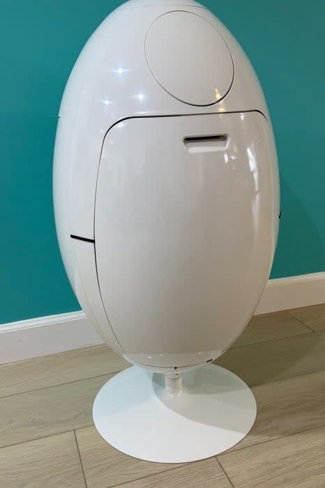 Egg-shaped trash can with a sleek white design, placed against a teal wall on a wooden floor