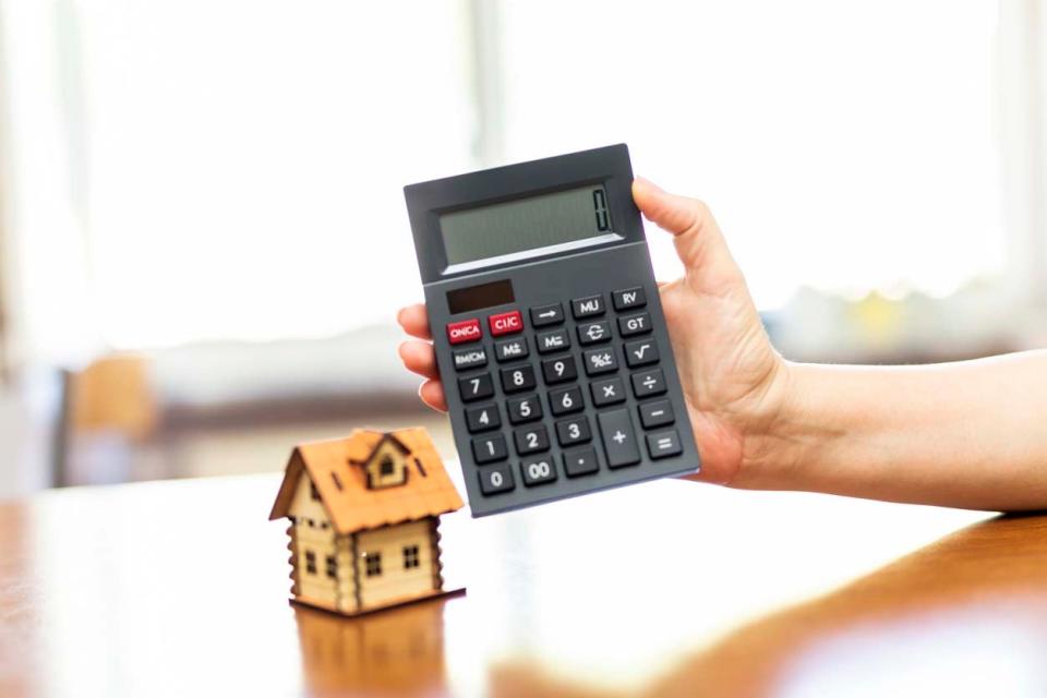 A hand holds up a grey calculator with a small model house in the background.