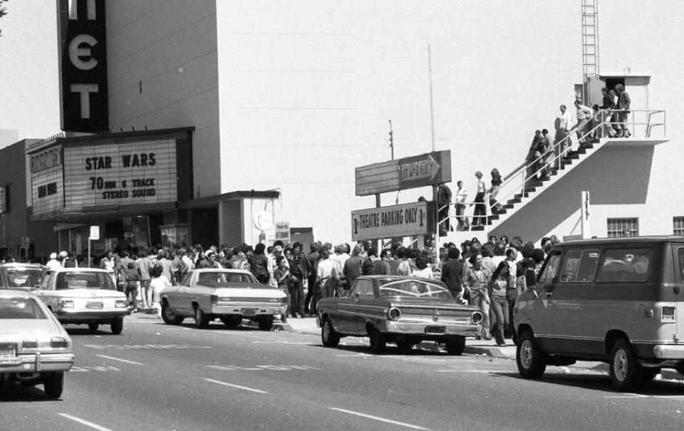 Crowd lining up outside a theater for "Star Wars" premiere, classic cars parked in front