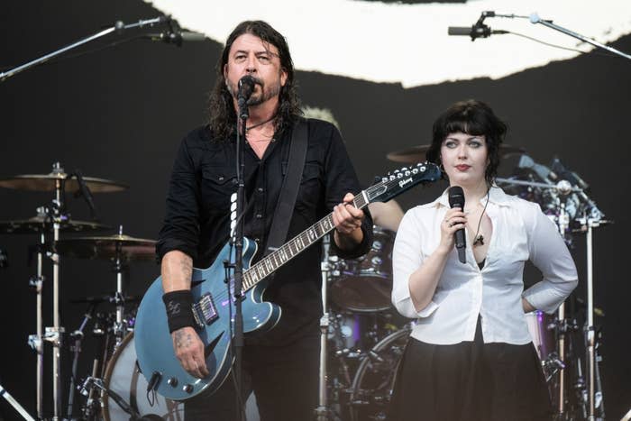 Dave Grohl, with a guitar, and Fiona Apple, with a microphone, perform on stage. Drums and other musical instruments are in the background