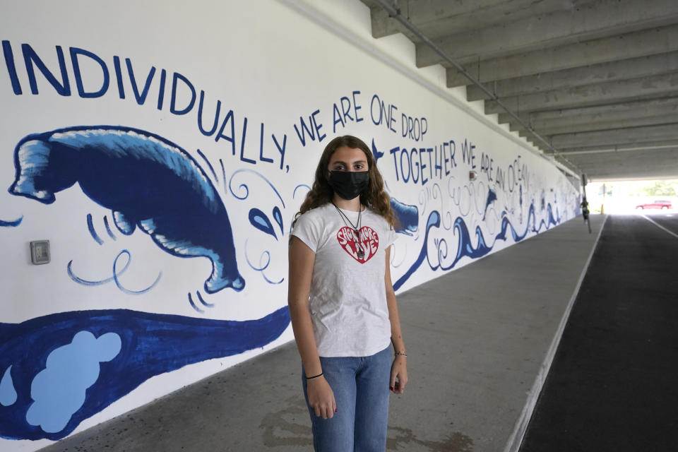 Holly Thorpe poses for a photo in front of a mural in the bus drop-off area at MAST Academy, that students painted with Carbon Dioxide (CO2) absorbing paint, Wednesday, Sept. 29, 2021, in Miami. Thorpe, 14, urged the Miami-Dade County Public Schools to considering replacing foul-smelling diesel school buses with electric vehicles. The school board voted this week to use a state grant to purchase up to 50 electric buses. Miami-Dade is joining a growing number of school districts transitioning from diesel to more environmentally-friendly electric school buses. (AP Photo/Wilfredo Lee)