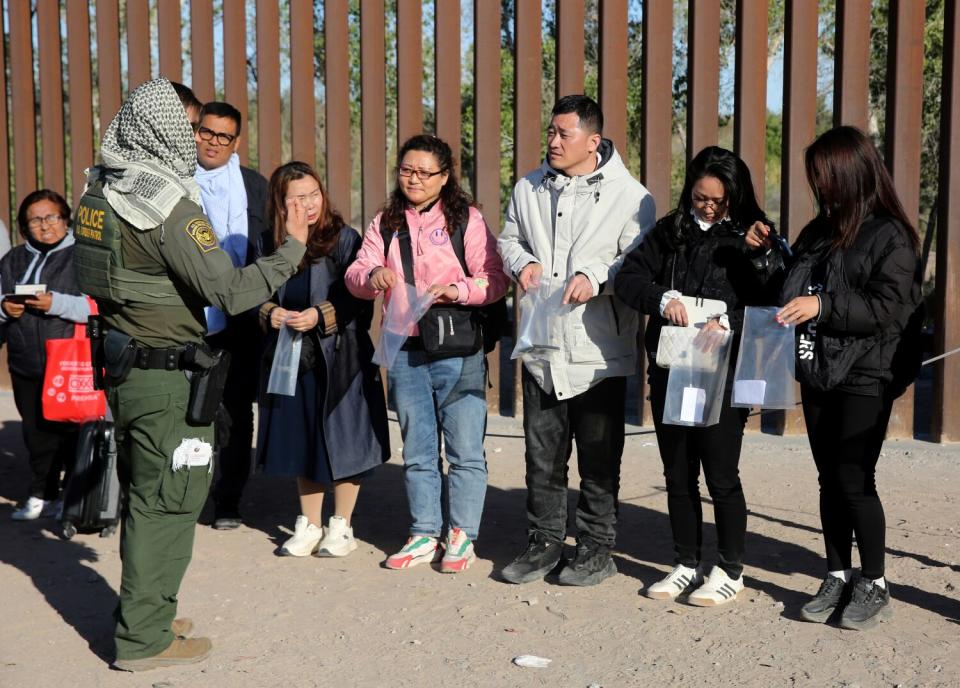 A group of migrants standing side by side next to a border fence, holding plastic bags and listening to a border agent