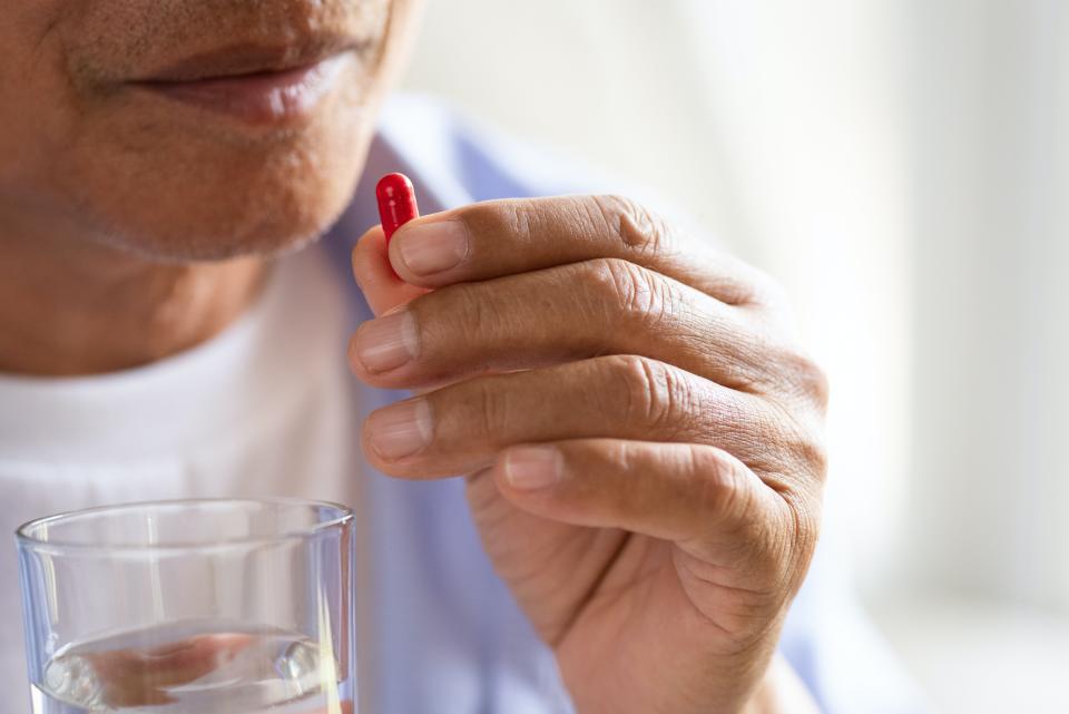 Un homme âgé porte un comprimé à sa bouche dans une main et un verre d’eau dans l’autre.