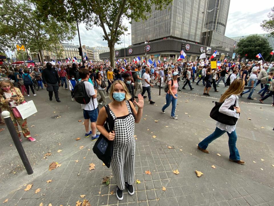 A pregnant woman wearing plaid overalls waves at the camera amid a crowd, wearing a mask.