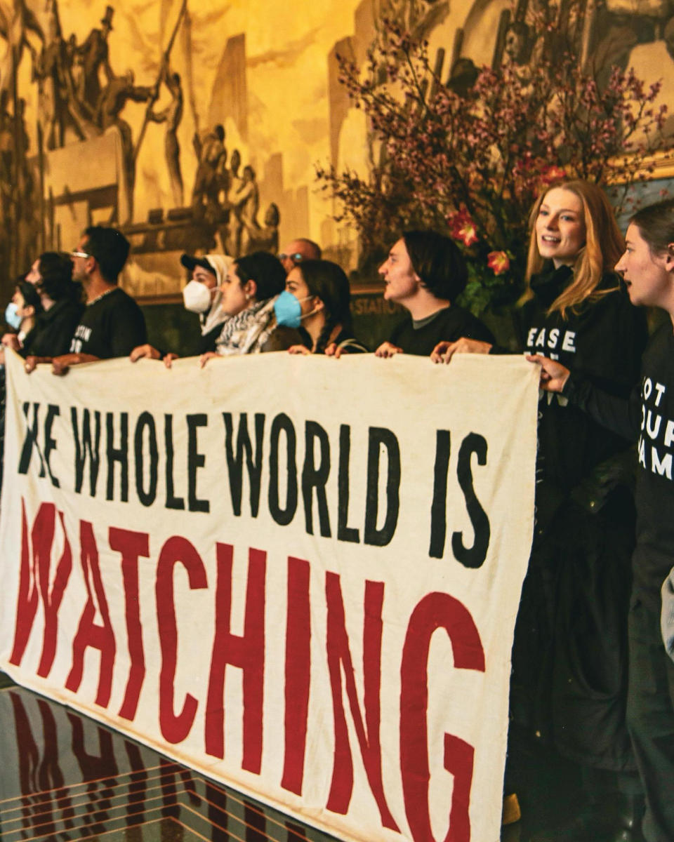 Group of individuals holding a banner reading "THE WHOLE WORLD IS WATCHING" at an event