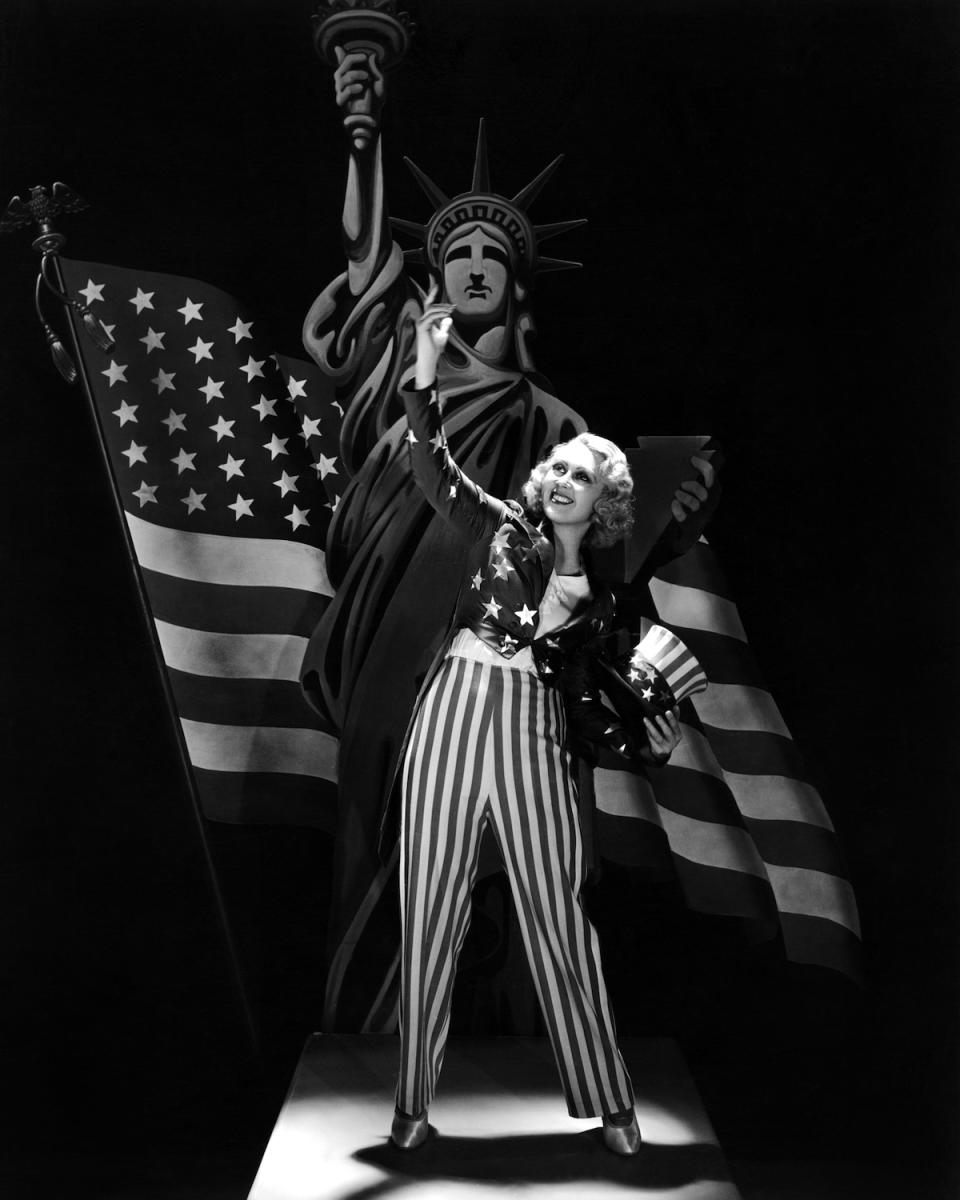 American actress Joan Blondell (1906 - 1978) in a patriotic pose, wearing an Uncle Sam outfit and standing in front of cutouts of the US flag and the Statue of Liberty, circa 1930