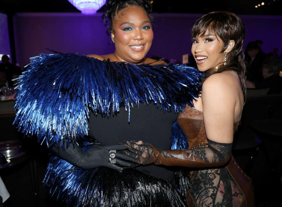 lizzo, wearing a black dress with blue grills and black gloves, holds hands and poses with cardi b, who wears a gold corset and black see through tights